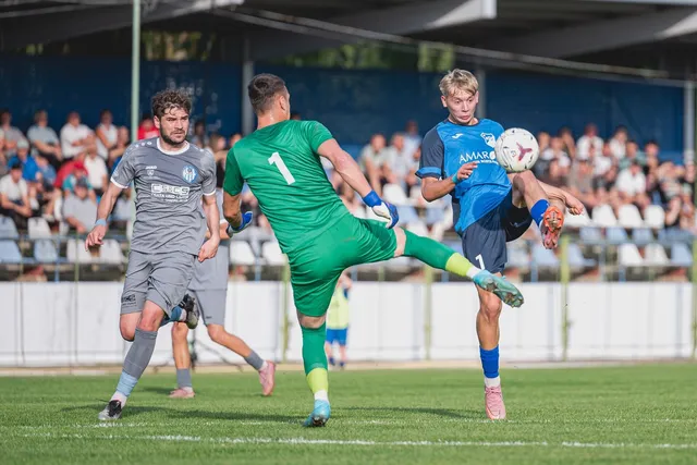Un jucător în echipament albastru lovește o minge de fotbal în aer, în timp ce un jucător în verde și unul în gri îl urmăresc pe teren.