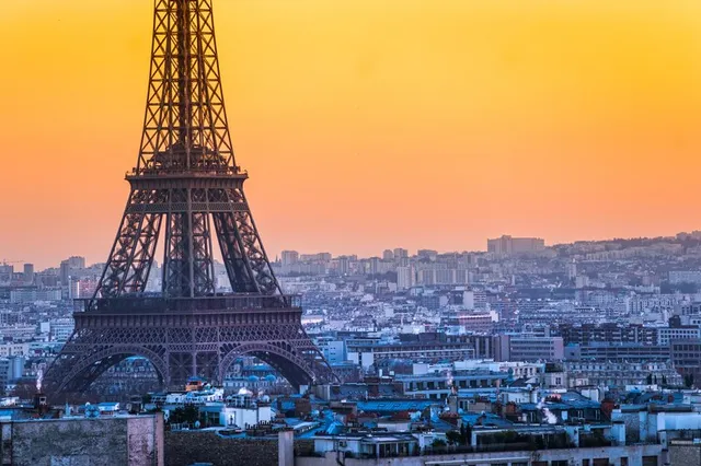 Turnul Eiffel se ridică deasupra unui peisaj urban dens sub un cer de culoare portocalie la apus.