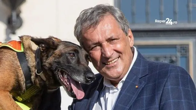 Un bărbat zâmbind poartă un sacou albastru și este alături de un câine cu harniş care are limba afară.