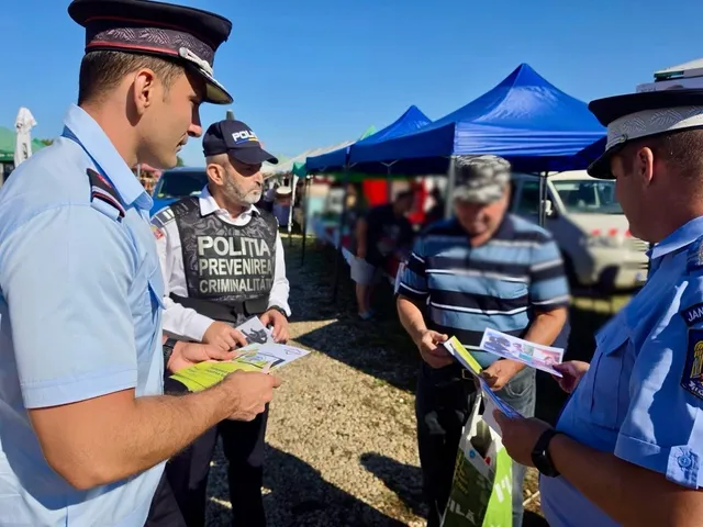 Trei ofițeri de poliție și jandarmi, doi dintre ei în prim plan, interacționează cu un civil, distribuind materiale informative în aer liber, sub un cer senin, cu corturi albastre în fundal.