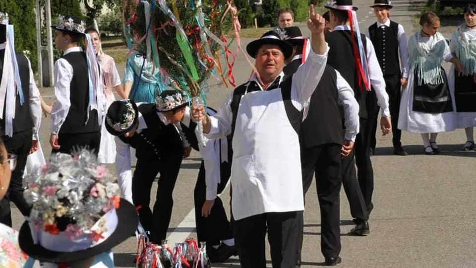 Un grup de persoane în costume tradiționale albe și negre merge pe o stradă, un bărbat ridicând mâna dreaptă în prim-plan.