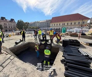 Oameni purtând veste de siguranță luminoase lucrează la o șantier cu conducte negre și o groapă deschisă în fața unor clădiri urbane.