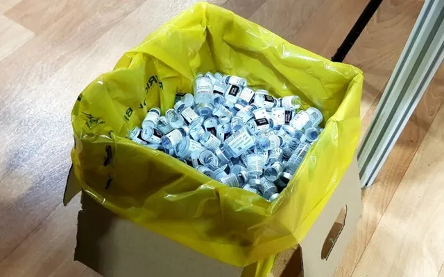 Un sac de plastic galben umplut cu numeroase flacoane mici de sticlă cu etichete albastre este așezat într-o cutie de carton pe un podea lemnoasă.