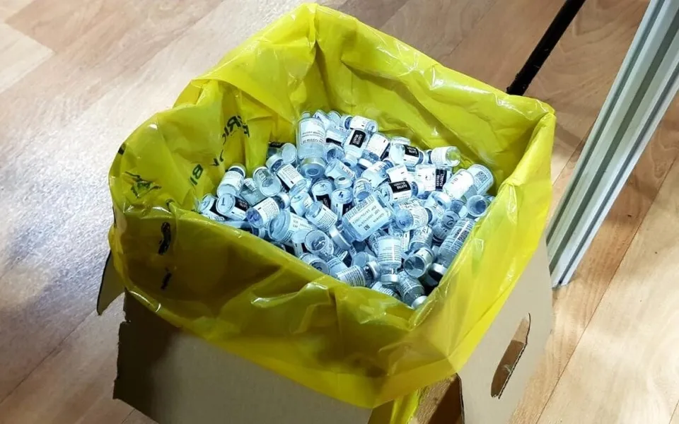 Un sac de plastic galben umplut cu numeroase flacoane mici de sticlă cu etichete albastre este așezat într-o cutie de carton pe un podea lemnoasă.