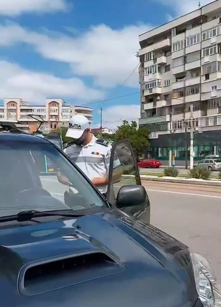 Un agent de poliție cu șapcă albă stă lângă portiera deschisă a unei mașini de culoare închisă pe un drum urban cu clădiri în fundal.