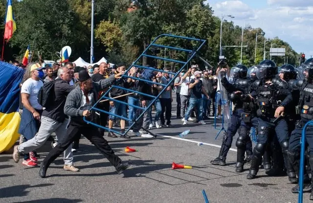 Un grup de persoane în haine civile se confruntă cu polițiști în echipament de protecție complet și scuturi pe o stradă asfaltată.