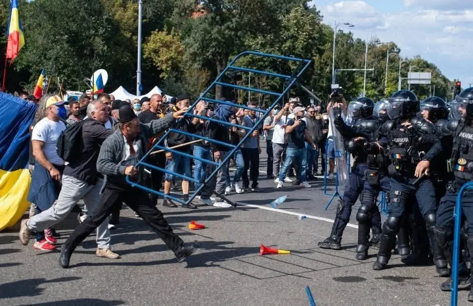 Un grup de persoane în haine civile se confruntă cu polițiști în echipament de protecție complet și scuturi pe o stradă asfaltată.