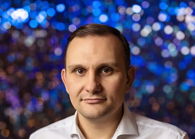 Un bărbat cu părul scurt și cămașă albă este poziționat în prim-plan în fața unui fundal albastru cu lumini difuze.
