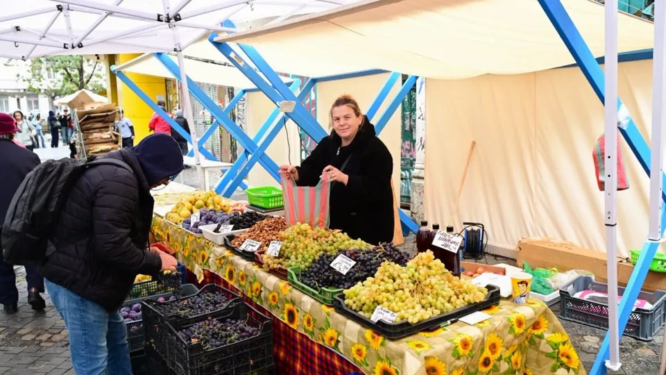 O femeie stă în spatele unui stand de fructe acoperit cu o față de masă cu modele de floarea-soarelui, în timp ce un bărbat privește spre stivă.