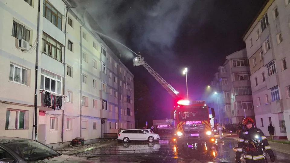 Un autospecială de pompieri cu lumină roșie este parcată pe o stradă între blocuri, sub o scară lungă extinsă spre acoperișul unei clădiri.