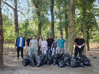 Un grup de persoane stă în rând în fața unor saci negri plini de deșeuri într-o zonă împădurită sub lumina soarelui.