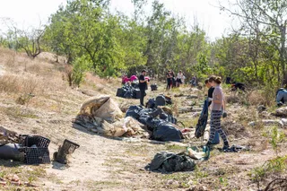 Oameni stau în apropierea unor saci negri de gunoi și resturi de materiale într-o zonă cu vegetație lemnoasă și teren nisipos.