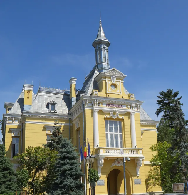 Clădire galbenă cu turn, acoperiș și coloane, înconjurată de arbori și steaguri, sub cer albastru.