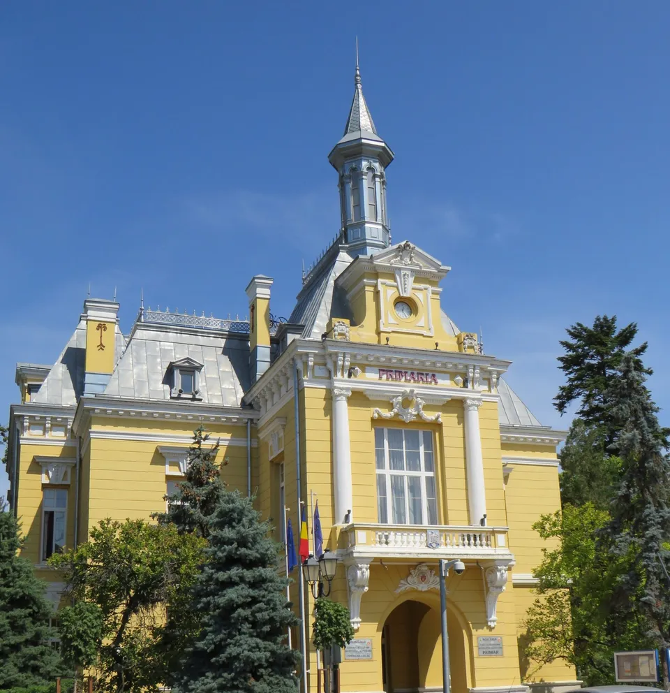 Clădire galbenă cu turn, acoperiș și coloane, înconjurată de arbori și steaguri, sub cer albastru.
