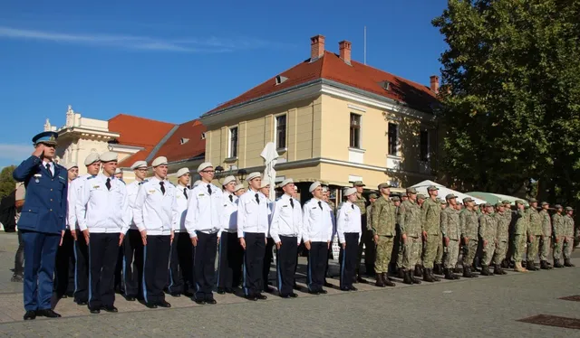 Un grup de persoane în uniforme albe și negre stă în rând, lângă un grup de persoane în uniforme de camuflaj, pe o suprafață pavată.