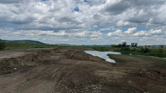 Un teren de pământ cu un șanț și o mică acumulare de apă sub un cer acoperit de nori albi și cenușii.