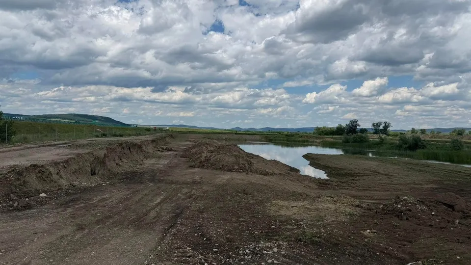 Un teren de pământ cu un șanț și o mică acumulare de apă sub un cer acoperit de nori albi și cenușii.