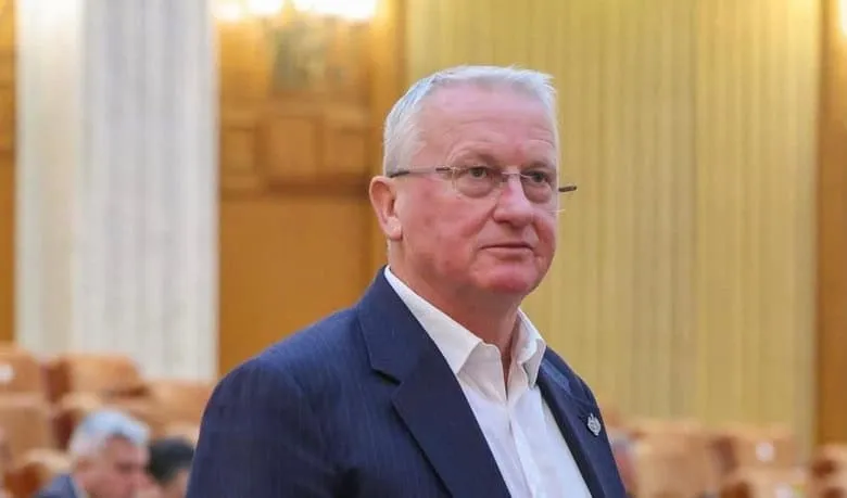 Un bărbat cu ochelari și sacou închis peste o cămașă albă stă în fața unui fundal cu panouri de lemn și scaune.