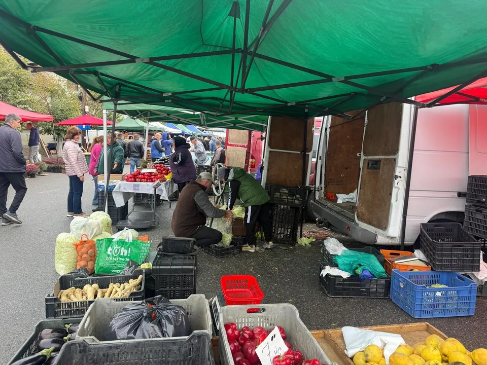 O piață acoperită cu un cort verde, cu vânzători și cumpărători în jurul de cutii cu fructe, legume și produse alimentare.
