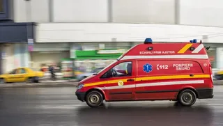 Un vehicul de serviciu roșu cu inscripții de ajutor medical și marcaje galbene circulă pe un drum asfaltat lângă clădiri.