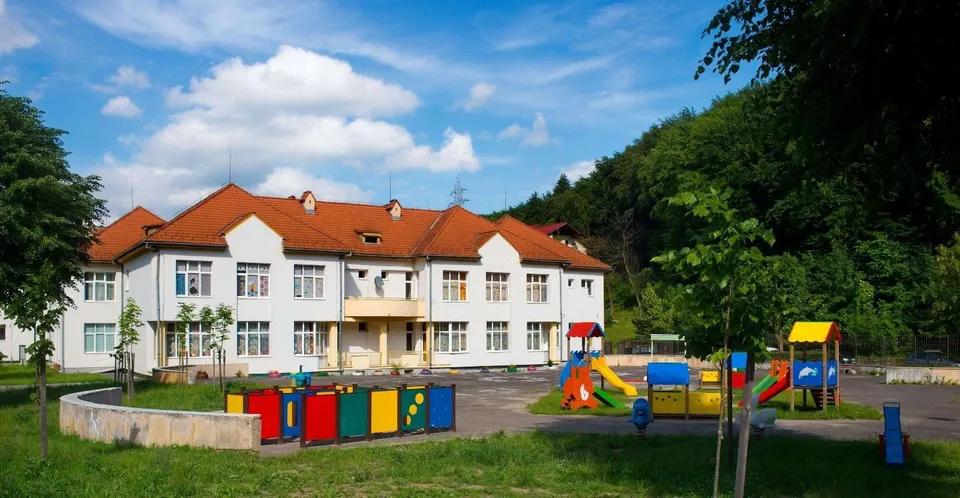 O clădire albă cu acoperiș roșu, cu un teren de joacă colorat în față, inconjurată de copaci și dealuri verzi.
