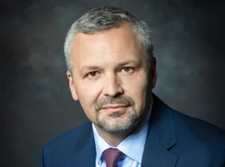 Un bărbat cu părul scurt și gri, purtând sacou albastru, cămașă deschisă la culoare și cravată roșie, privește spre cameră pe un fundal întunecat.