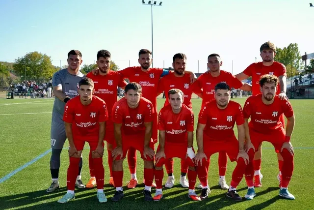 O echipă de fotbaliști îmbrăcați în echipamente roșii pozează pe un teren de fotbal sub cerul senin.