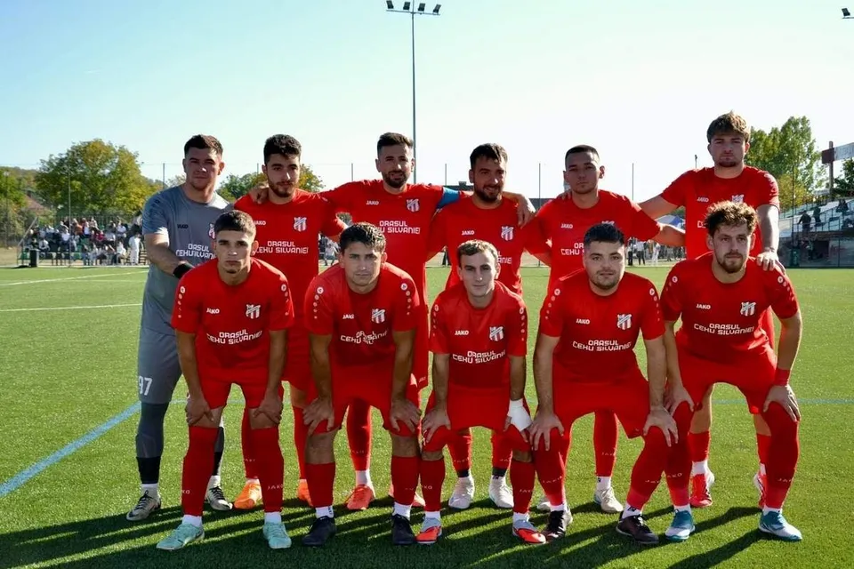 O echipă de fotbaliști îmbrăcați în echipamente roșii pozează pe un teren de fotbal sub cerul senin.
