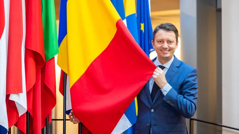 Un bărbat într-un costum albastru ține o steag tricolor galben, roșu și albastru în fața mai multor steaguri colorate.