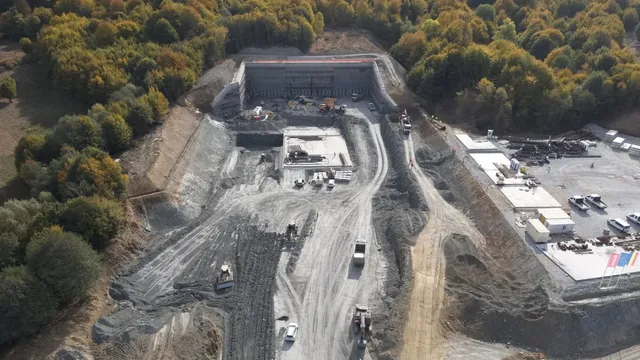 O vedere aeriană a unui șantier de construcții cu utilaje, drumuri de pământ și o intrare de tunel înconjurată de vegetație densă.