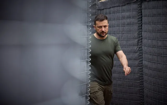 Un bărbat cu barbă și tricou verde merge lângă un gard de sârmă și un panou textil gri.