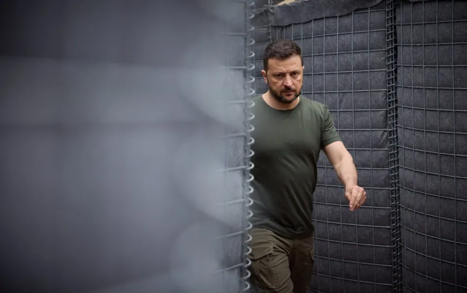 Un bărbat cu barbă și tricou verde merge lângă un gard de sârmă și un panou textil gri.