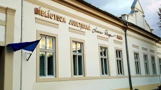 O clădire albă cu ferestre înalte și un steag albastru prăbușit pe o parte, având inscripția Biblioteca Județeană Alba pe fațadă.