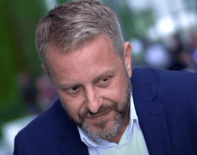 Un bărbat cu păr și barbă grizonante, în sacou albastru și cămașă deschisă, privește în jos, pe un fundal verde neclar.