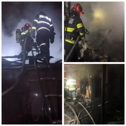 Pompieri în echipament de protecție și cu echipament de respirație lucrează noaptea lângă structuri de lemn în presence de fum și furtunuri de apă.