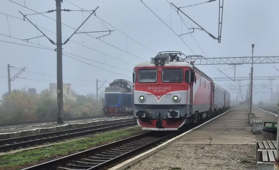 Un tren roșu și gri circulă pe un drum feroviar lângă un peron, sub linii electrice suspendate într-o zi cu ceață.