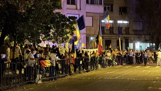 O mulțime de oameni stă în spatele unor bariere metalice pe un drum, lângă un clădire cu mai multe steaguri colorate afișate.