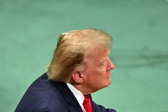 Un bărbat cu păr blond, purtând un costum închis la culoare, o cămașă albă și o cravată roșie, este surprins în profil pe un fundal verde.