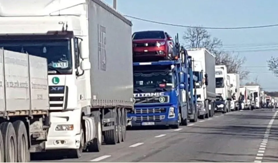 Mai multe camioane de marfă sunt parcate în rând pe o șosea asfaltată sub un cer senin.