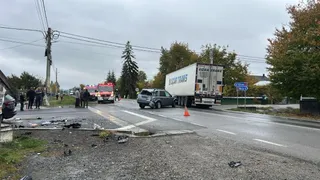 Un autocamion alb și un vehicul gri sunt pe un drum asfaltat lângă resturi împrăștiate pe marginea drumului sub un cer înnorat.