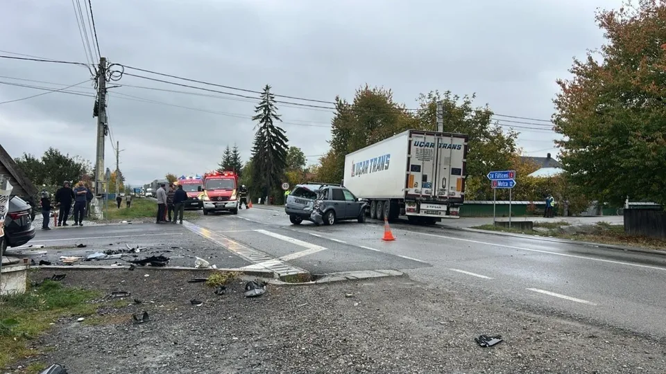 Un autocamion alb și un vehicul gri sunt pe un drum asfaltat lângă resturi împrăștiate pe marginea drumului sub un cer înnorat.