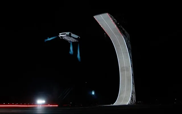 Un vehicul alb cu jeturi albastre emise de sub acesta zboară pe fundalul unui cer negru lângă o rampă curbată de beton.