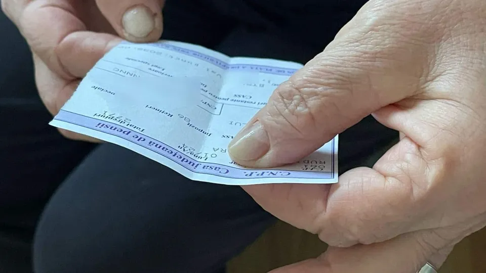 Două mâini țin și deschid un document de format mic cu text și tabele imprimate pe fundal albastru și alb.