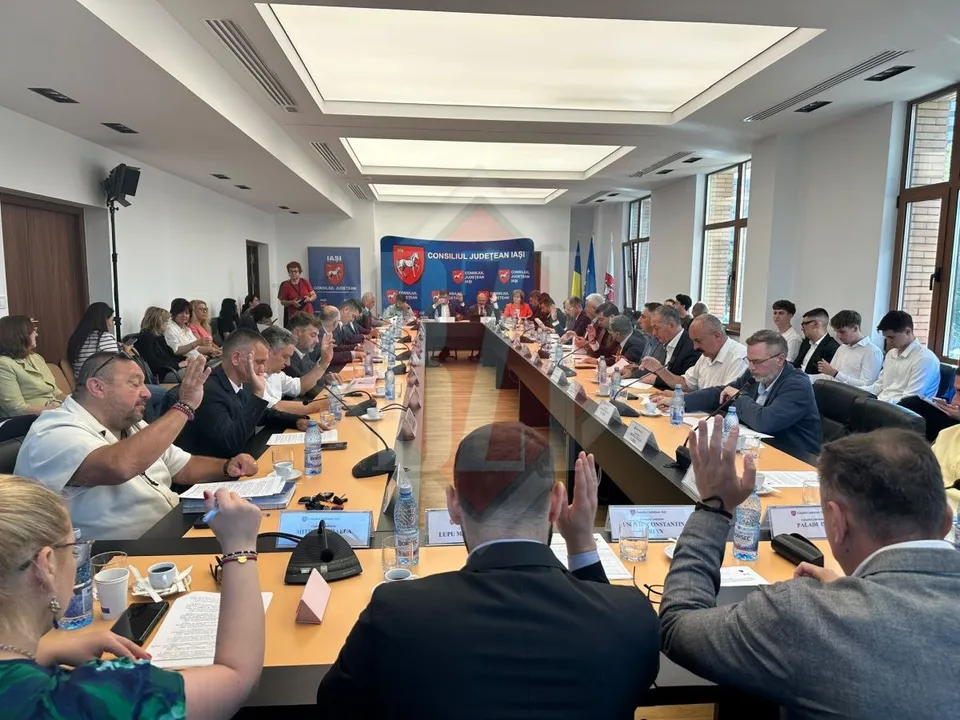 O grupă de persoane stă la mese lungi în fața unui panou albastru într-o sală de ședințe cu ferestre înalte.