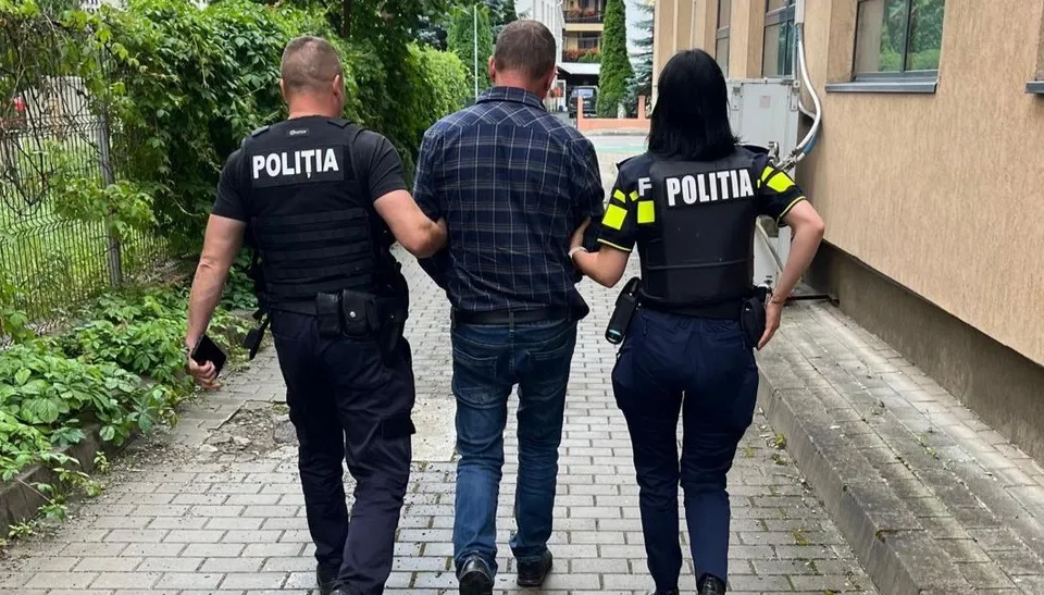 Doi polițiști în uniformă negru și galben conduc un bărbat cu cămașă în carouri și pantaloni albaștri pe un drum pavat.