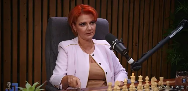 O femeie cu părul roșu, purtând o sacou roz deschis, stă la o masă lângă un microfon și un set de piese de șah.