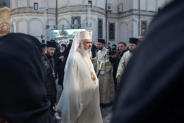 Un bărbat cu barbă și pălărie albă, purtând o mantie albă cu un medalion aurit, merge printre alți bărbați îmbrăcați în haine negre și albe.