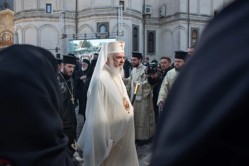 Un bărbat cu barbă și pălărie albă, purtând o mantie albă cu un medalion aurit, merge printre alți bărbați îmbrăcați în haine negre și albe.