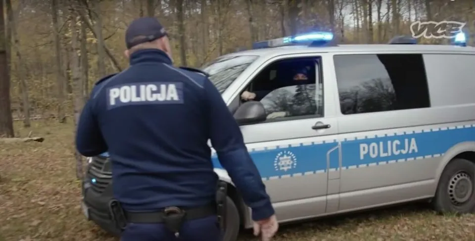 Un polițist cu un tricou albastru pe care scrie POLICJA stă lângă un vehicul alb cu marcaje și lumini albastre de poliție într-o zonă împădurită.