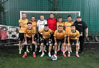 Un grup de zece bărbați pozează pe un teren de fotbal cu un balon alb și albastru în prim-plan, în fața unei porți de fotbal.